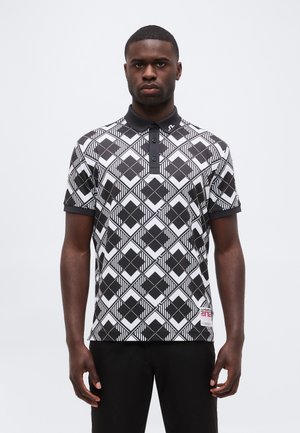 Homme debout face à l'avant, portant un polo à motif géométrique noir et blanc et un pantalon noir sur un fond clair uni.