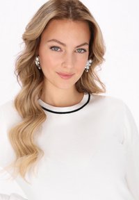 Pulover alb cu guler rotund și margini negre, dintr-un material texturat, și părul ondulat blond deschis. Modelul poartă cercei mari cu accente argintii.