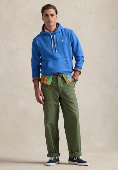 Jaunas vyras stovintis, dėvintis mėlynį džemperį per daudziaspalvį marškinį, žalius kelnes ir mėlynas sportbačius, priešais paprastą smėlio spalvos foną.