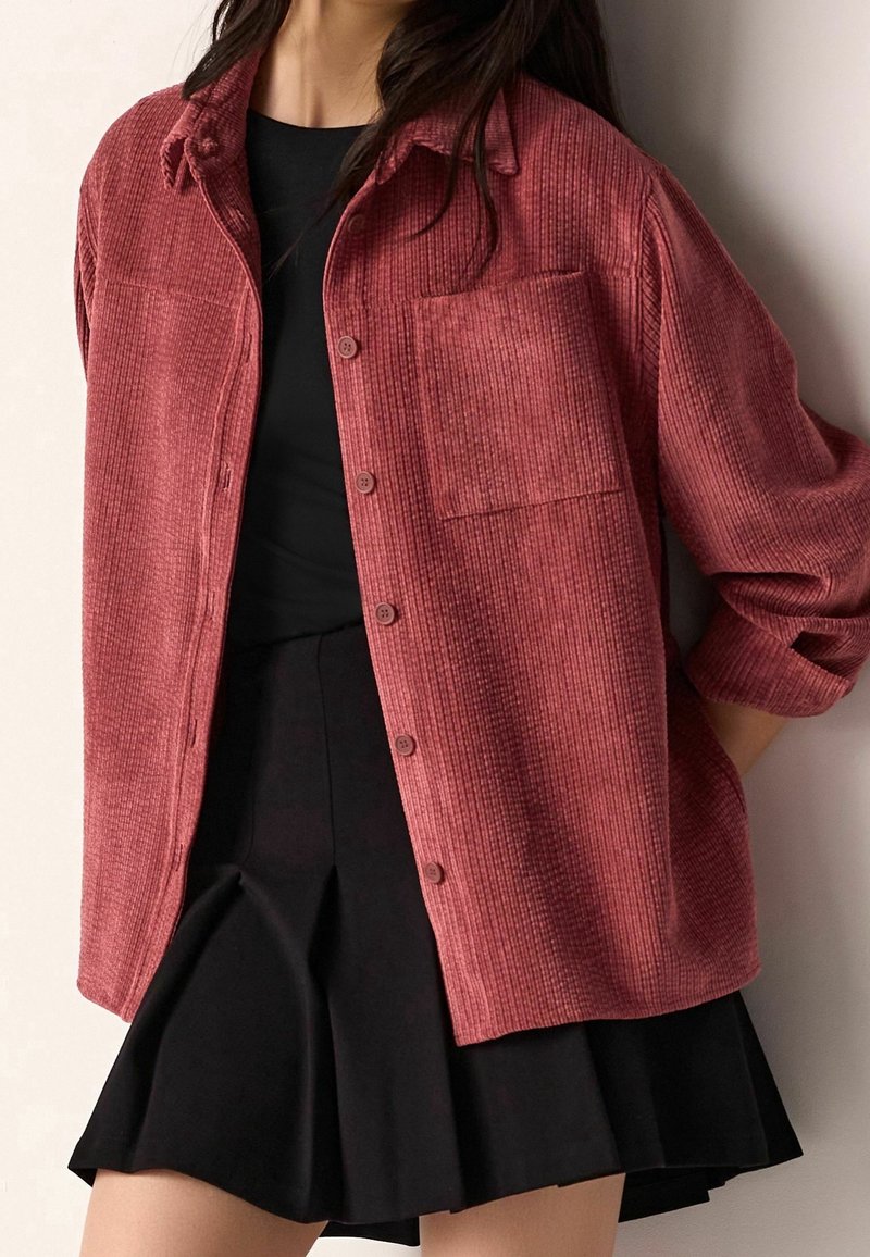 Femme portant une chemise en velours côtelé rouge boutonnée sur un haut noir et une jupe plissée noire, debout avec une main derrière le dos.