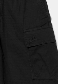 Sort stof med tekstureret gittermønster, der har en syet cargolomme og broderet "SWTZ"-logo på lommens klap.