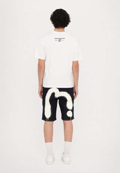 T-shirt en coton blanc avec du texte noir au dos, associée à un short en denim foncé présentant un grand design de point d'interrogation pulvérisé en blanc.