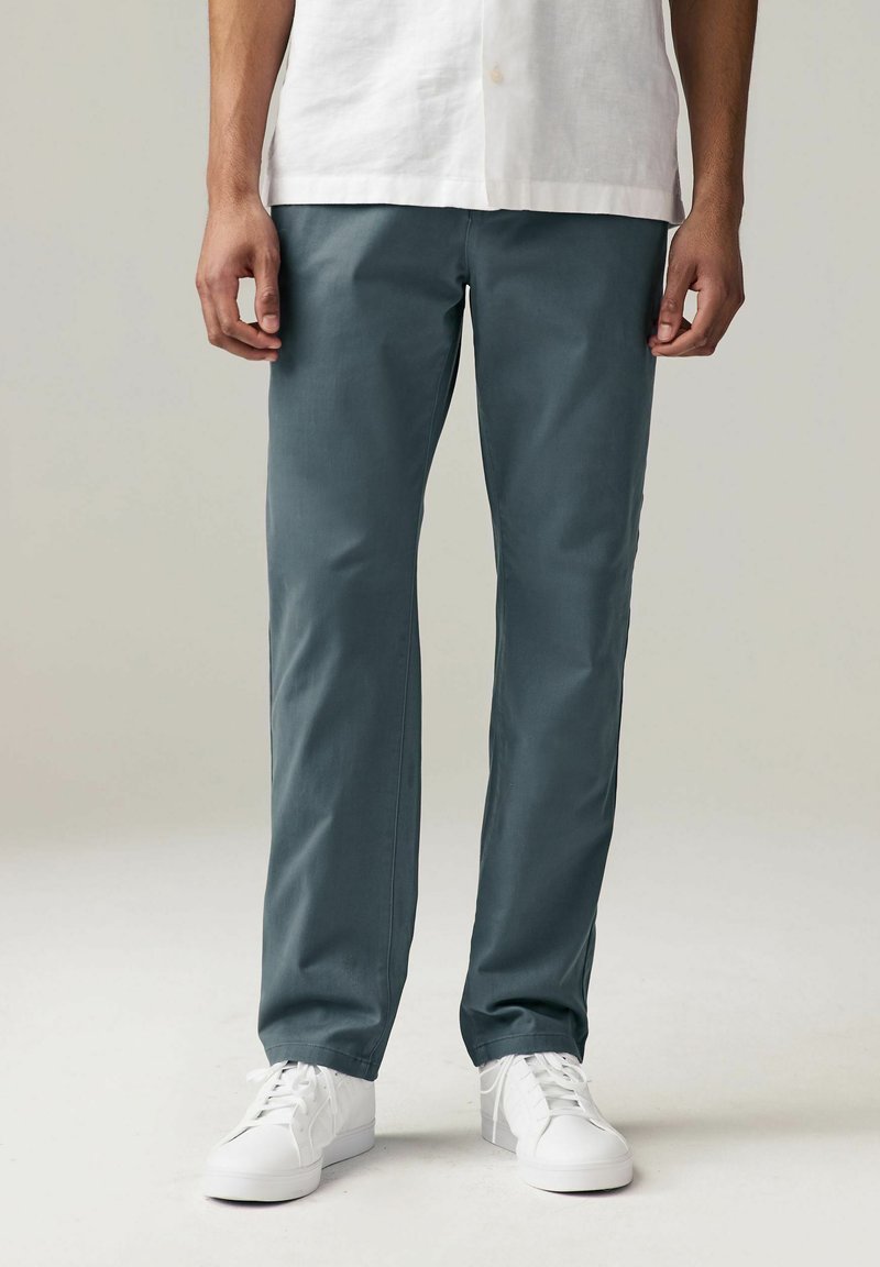 Pantaloni slim-fit in un morbido tessuto teal, con una superficie liscia e gambe dritte, abbinati a sneaker bianche per un look casual.