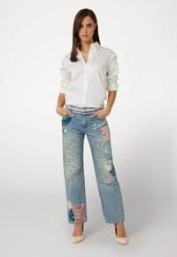 Balta mygtukų marškiniai ir mėlyni dėvėti džinsai su įvairiomis grafikos aplikacijomis ir siuvinėjimais. Šviesios spalvos aukštakulniai batai papildo aprangą.