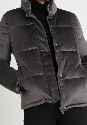 Ciemnoszara aksamitna kurtka puchowa z przednim zamkiem błyskawicznym i zatrzaskami, noszona na czarnym golfie, z rękami w bocznych kieszeniach.