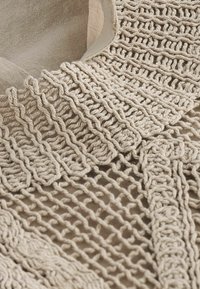 Tissu tricoté beige avec des motifs complexes de boucles torsadées et ouvertes, montrant la texture et les détails du fil tissé en gros plan.