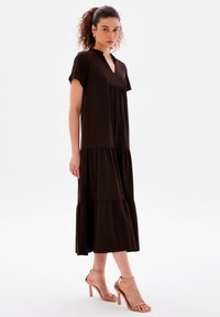 Braunes, knielanges Kleid mit einem geschichteten Design, kurzen Ärmeln, einem Schlüsselloch-Ausschnitt und glatter Textur; kombiniert mit nude-farbenen, geflochtenen High-Heel-Sandalen.