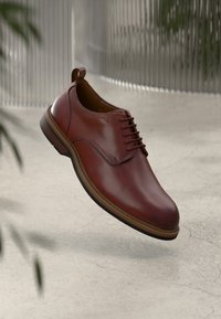 Brown leather lace-up dress shoe with a pull tab, floating above a light-colored textured floor and blurred background.