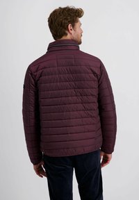 Man die met zijn rug naar de camera staat, gekleed in een gewatteerde, donkerpaarse jas met horizontale stiksels en zwarte broek, tegen een effen lichte achtergrond.