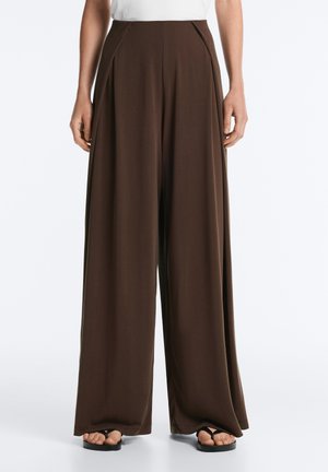 Vue de face d'une personne portant un pantalon ample, taille haute, marron foncé, à jambes larges et des sandales noires, debout sur un fond blanc.