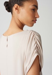 Zachte beige top met een ontspannen pasvorm, korte mouwen met geplooide details en een kleine knoopsluiting bij de achterkant van de hals.