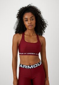 Mujer con cabello rizado que lleva un sujetador deportivo y leggings de Under Armour en color burdeos a juego, de pie contra un fondo claro y sencillo.