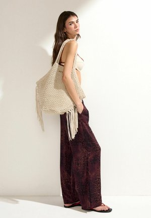 Ung kvinde iført mørke mønstrede vidde bukser og sandaler, bærende en stor cremefarvet strikket taske med frynser over skulderen.