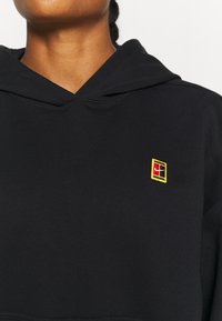 Schwarzer Hoodie aus weichem Stoff mit einem gelb-roten gestickten Logo auf der Brust. Strukturiertes Kapuzendesign mit Rippdetails.
