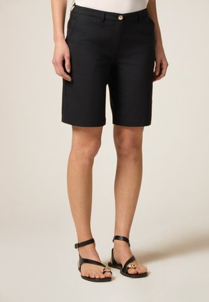 Shorts neri su misura con una texture liscia, chiusura con zipper e bottone, tasche laterali e cuciture a contrasto. Abbinati a sandali neri.