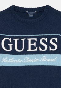 Mornarska pletena jopica s krožnim ovratnikom; vsebuje belo črkovanje "GUESS" in svetlo modro akcentno črto, z "Authentic Denim Brand" v kurzivi.
