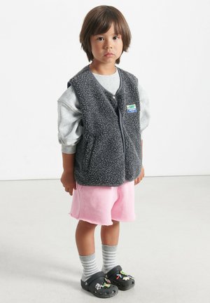 Grå fleecet väst med en strukturerad yta, rosa shorts, randiga grå och vita strumpor, samt svarta crocs med färgglada dekorationer.