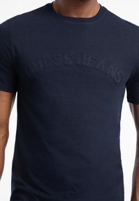 Námořnická modrá bavlněná tričko s vyvýšeným tmavým nápisem "GUESS JEANS" přes hrudník, s kulatým výstřihem a krátkými rukávy.