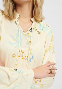 Krémová blůza s květinovými vzory v modré, žluté a černé barvě. Obsahuje výstřih do V, detail s knoflíky a široké rukávy se shromážděnými akcenty.