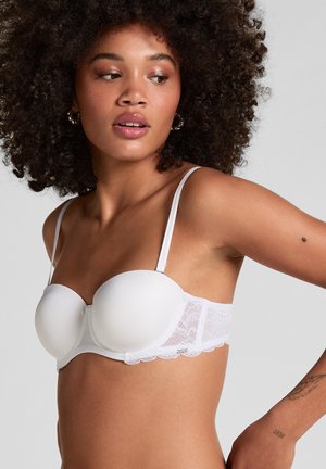 Vrouw met krullend haar die een witte strapless beha met kanten details draagt, kijkt naar de zijkant tegen een effen lichtgrijze achtergrond.