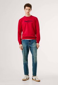Sweatshirt vermelho com logótipo azul-marinho, combinado com jeans azul claro. A modelo usa ténis brancos com detalhes pretos. Ajuste casual e estilo moderno.
