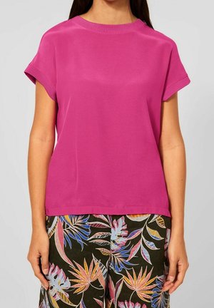 Fuchsiafarbenes Kurzarmoberteil mit rundem Ausschnitt und strukturiertem Rippbesatz. Kombiniert mit gemusterten Blumenhosen in lebendigen Farben auf dunklem Hintergrund.