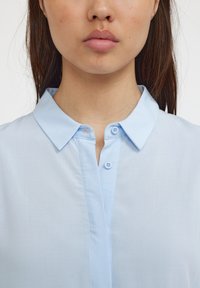 Hele sinine krae-särk, millel on nööbitud plisseeritud, lühikesed varrukad ja sile koe tekstuur. Omab kahte nuppu ja puhast disaini.