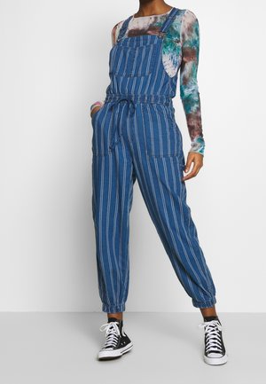 Person iført blå og hvide denim-alls med striber med elastiske manchetter og en flerfarvet langærmet tie-dye-shirt, hænderne i lommerne.