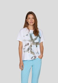 Weißes Baumwoll-T-Shirt mit abstraktem blau-grünem Grafikdruck, lässiger Schnitt, kurze Ärmel und runder Ausschnitt, kombiniert mit hellblauen Hosen.