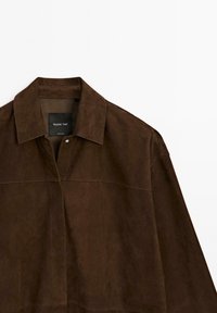 Brown suede jacket with a collared neck, featuring a label, intricately stitched seams, and a smooth texture against a white background.