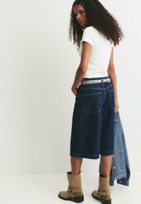 Donna dai capelli ricci indossa un top bianco a maniche corte, pantaloni corti di jeans scuri al ginocchio, stivali color cammello e tiene in mano una giacca di jeans in uno studio bianco.
