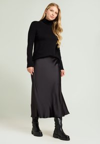 Schwarzer Rollkragenpullover und ein schwarzer Satin-Maxi-Rock, kombiniert mit knielangen schwarzen Lederstiefeln, vor einem hellen Hintergrund.