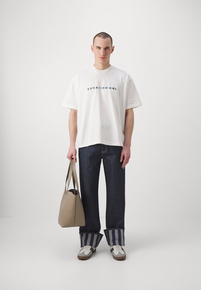 T-shirt en coton blanc avec du texte, jean bleu foncé avec des poignets rayés, sac fourre-tout beige et baskets blanches avec des accents marron.