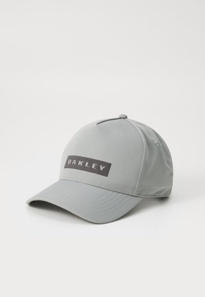 Pilkas beisbolo kepurė, pagaminta iš lengvos medžiagos, su juodu stačiu kampu pleistru, ant kurio užrašyta "OAKLEY" baltomis stora raidėmis.