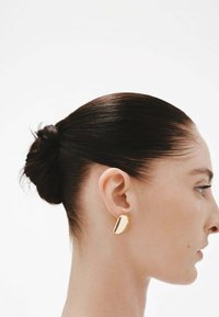 Pendiente geométrico dorado sobre un tono de piel suave. El cabello está recogido en un moño elegante. La vista de perfil muestra claramente la oreja y el pendiente contra un fondo blanco.