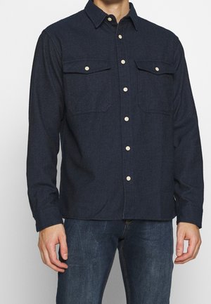 Homme portant une chemise bleu marine foncé à boutons avec poches à rabat sur la poitrine et un jean bleu sur un fond clair uni.