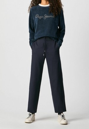 Personne portant un sweat Pepe Jeans bleu foncé, un pantalon large bleu marine à fines rayures et des baskets blanches sur un fond uni.