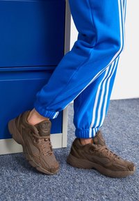 Brown mesh sneakers with laces and a textured finish, featuring a reinforced toe and heel. Paired with blue track pants with white stripes.