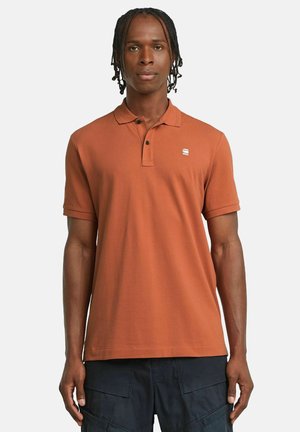 Brunorange polo skjorta med en tre-knapplåsning, korta ärmar och en liten vit logotyp på bröstet. Slät, bomullsblandad textur.