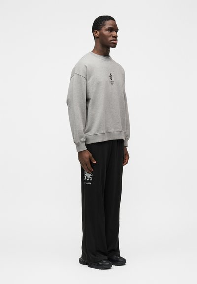 Hanorac gri cu decolteu rotund, asortat cu pantaloni largi negri, cu un detaliu grafic alb. Modelul este în poziție de profil.