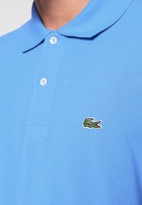 Polo bleu clair en tissu texturé, avec un col et un petit logo crocodile vert sur la poitrine gauche.