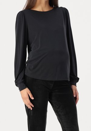Femme portant un chemisier noir à manches longues avec poignets froncés et pantalon noir, vue des épaules jusqu'au milieu des cuisses sur fond blanc.