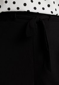 Czarny pasek materiału z wiązanym kokardą z przodu, w połączeniu z białą bluzką w czarne kropki. Gładka faktura.