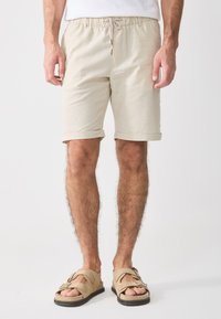 Beige cotton shorts with an elastic waistband and drawstring, featuring turned-up cuffs. Paired with tan sandals.