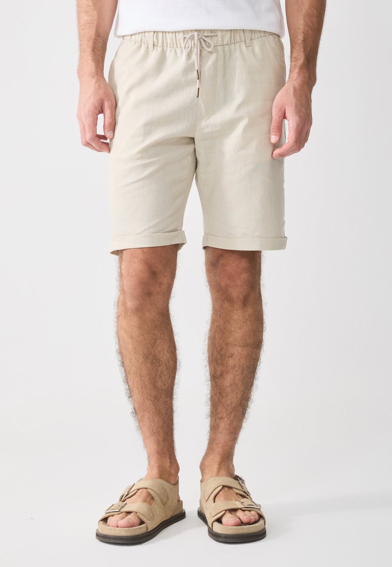 Beige cotton shorts with an elastic waistband and drawstring, featuring turned-up cuffs. Paired with tan sandals.