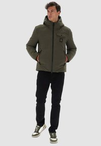 Giacca a vento verde oliva con cappuccio, zip anteriore e tasca sul petto. Abbinata a pantaloni neri e sneakers con dettagli verdi.