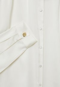 Blusa in seta bianca con patta di bottoni, maniche lunghe con dettaglio al polsino e accentuazione in tono oro, texture liscia con leggera lucentezza.