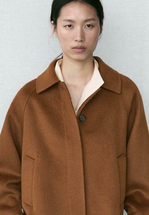 Hnědý vlněný kabát s klopovým límcem, jedním černým knoflíkem a bočními kapsami. Kabát má měkkou texturu a volný střih.