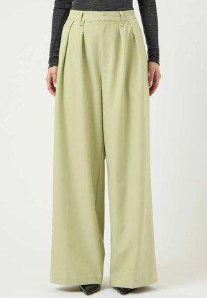 Mujer con pantalones verde claro de tiro alto y pierna ancha combinados con una blusa de manga larga gris oscuro y zapatos negros puntiagudos.