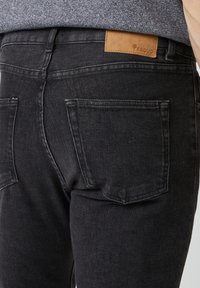 Vista trasera de una persona que lleva puestos jeans gris oscuro con dos bolsillos traseros y una etiqueta de parche de cuero marrón en la cintura.